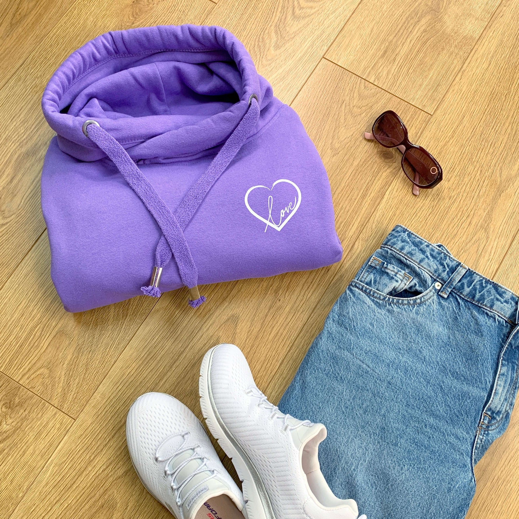 Folded lavender purple relaxed fit Love heart cross cowl neck casual fashion hoodie. Cotton Polyster blend relaxed fit unisex oversized hoodie design has love heart. White heart to upper left of the chest with word Love in white handwriting font nestled within the heart. Wooden floor sunglasses blue jeans and white trainers