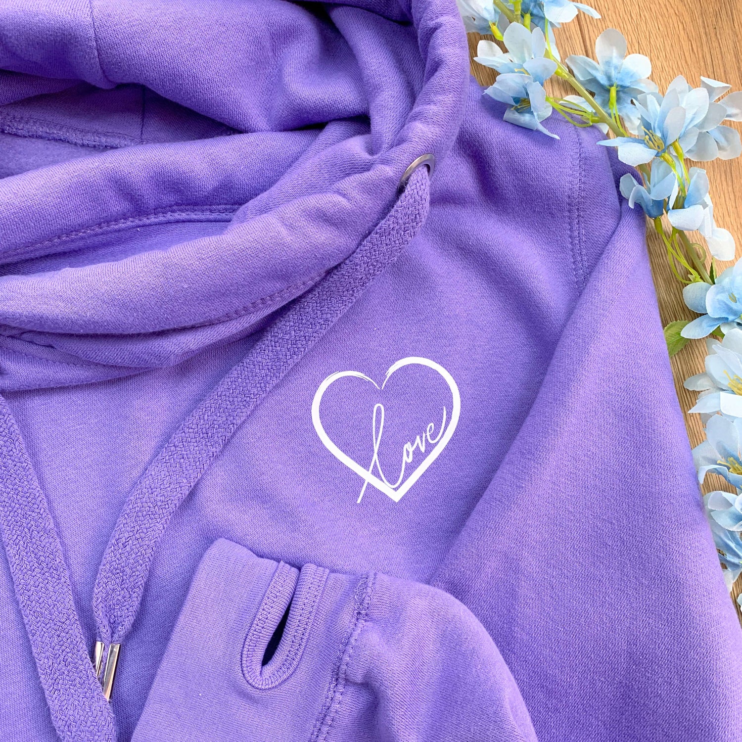 Close up photo of purple lavender unisex Love heart cross cowl neck hoodie. Cotton Polyester blend oversized casual style hoodie design has small love heart to upper left chest. White heart with word Love in white handwriting font nestled within the heart. Drawstrings with metal toggles to ends and thumb holes to long sleeve arm cuffs. Blue spring decorative flowers to right of photo resting next to top on a wooden floor