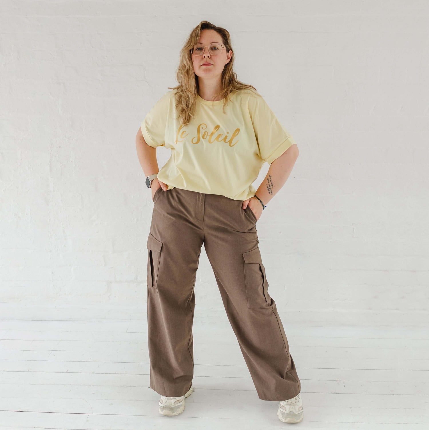 Woman with blonde hair and glasses, wearing butter light yellow oversized t-shirt with loose dolman sleeves with rolled tacked cuff and round neckline loosely tucked into taupe cargo trousers.. Ladies t shirt has the word design Le Soleil across the chest in matt metallic gold script font.