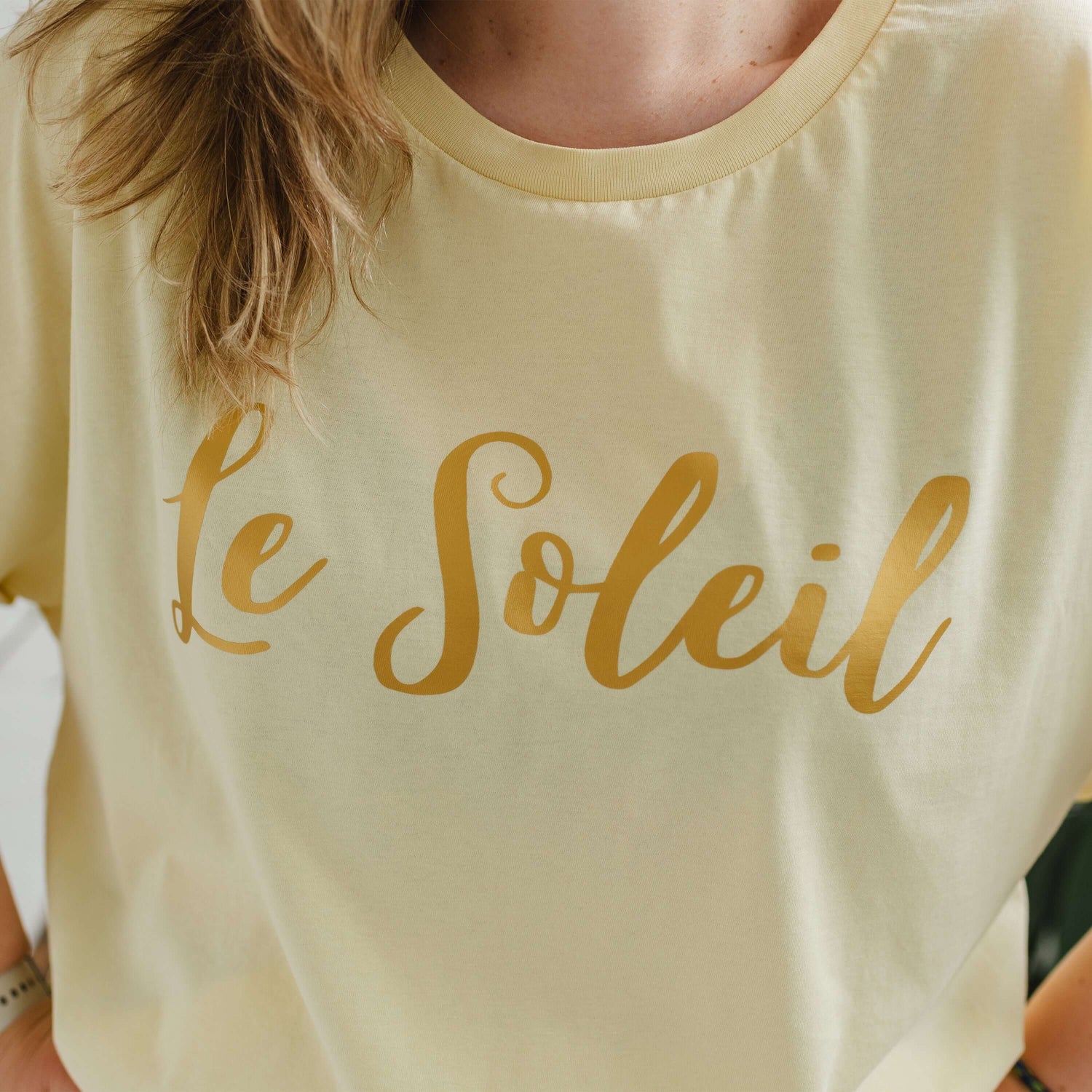 Close up of woman with blonde hair wearing butter light yellow oversized t-shirt with loose dolman sleeves with rolled tacked cuff and round neckline. Ladies t shirt has the word design Le Soleil across the chest in matt metallic gold script font.