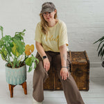 Woman sitting on a wooden weathered chest. Blonde hair and glasses, wearing butter light yellow oversized t-shirt with loose dolman sleeves with rolled tacked cuff and round neckline loosely tucked into taupe cargo trousers.. Ladies t shirt has the word design Le Soleil across the chest in matt metallic gold script font. Woman wears taupe casual baseball cap