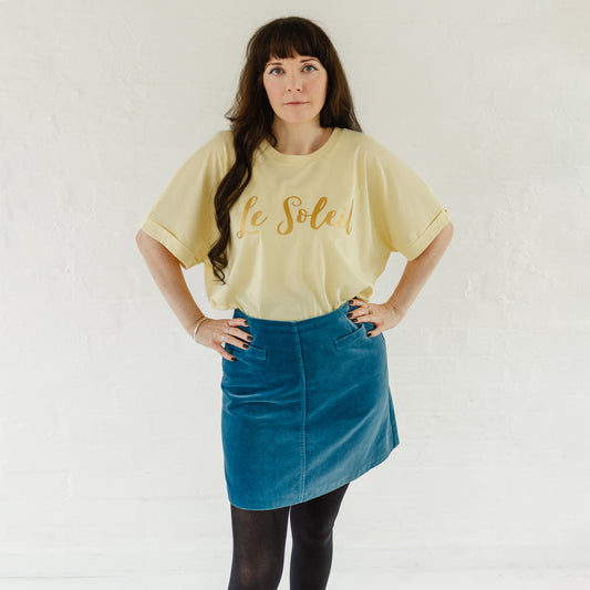 Woman wearing butter light yellow oversized t-shirt with loose dolman sleeves with rolled tacked cuff and round neckline tucked into a velvet mid blue a line skirt. Ladies t shirt has the word design Le Soleil across the chest in matt metallic gold script font. 