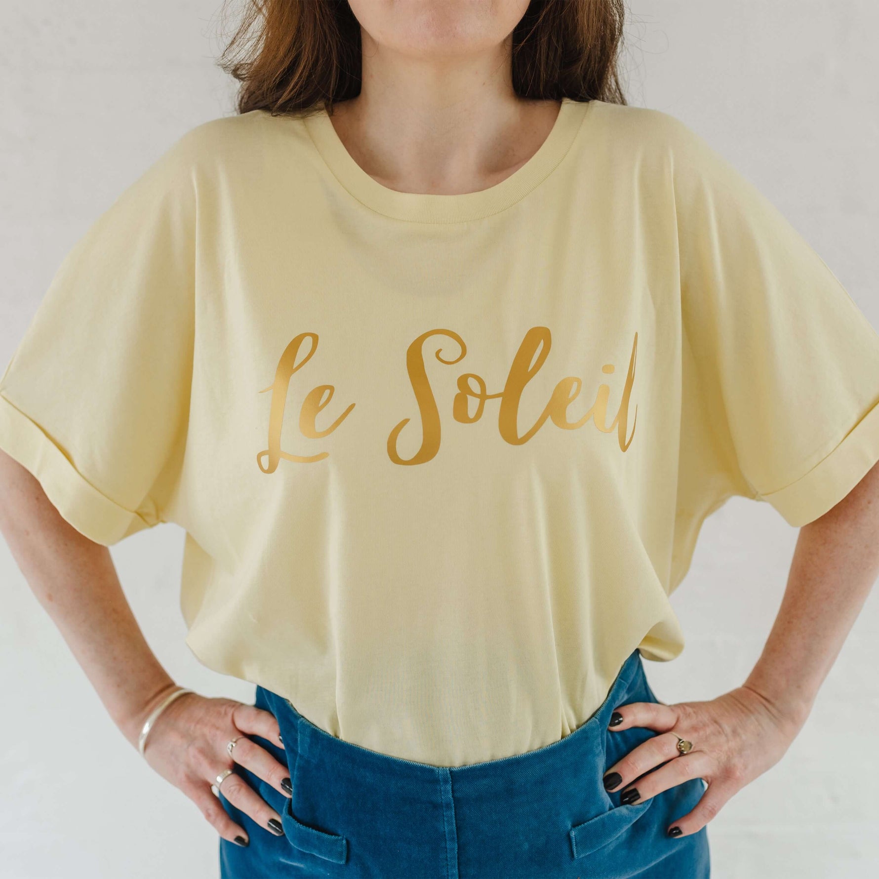 Close up of woman wearing butter light yellow oversized t-shirt with loose dolman sleeves with rolled tacked cuff and round neckline tucked into a velvet mid blue a line skirt. Ladies t shirt has the word design Le Soleil across the chest in matt metallic gold script font.