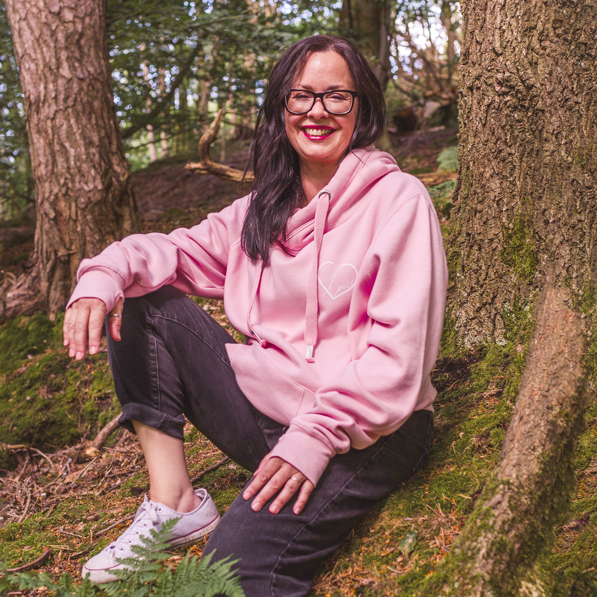 Ladies casual hoodie. Woman sitting smiling in wooden area wears baby pink womens hoodie Love heart cross cowl neck casual fashion hoodie. Cotton Polyester blend relaxed fit unisex oversized style hoodie design has love heart. Whitert to upper left of the chest with word Love in white script font nestled within the heart, paired with black straight leg jeans and trainers