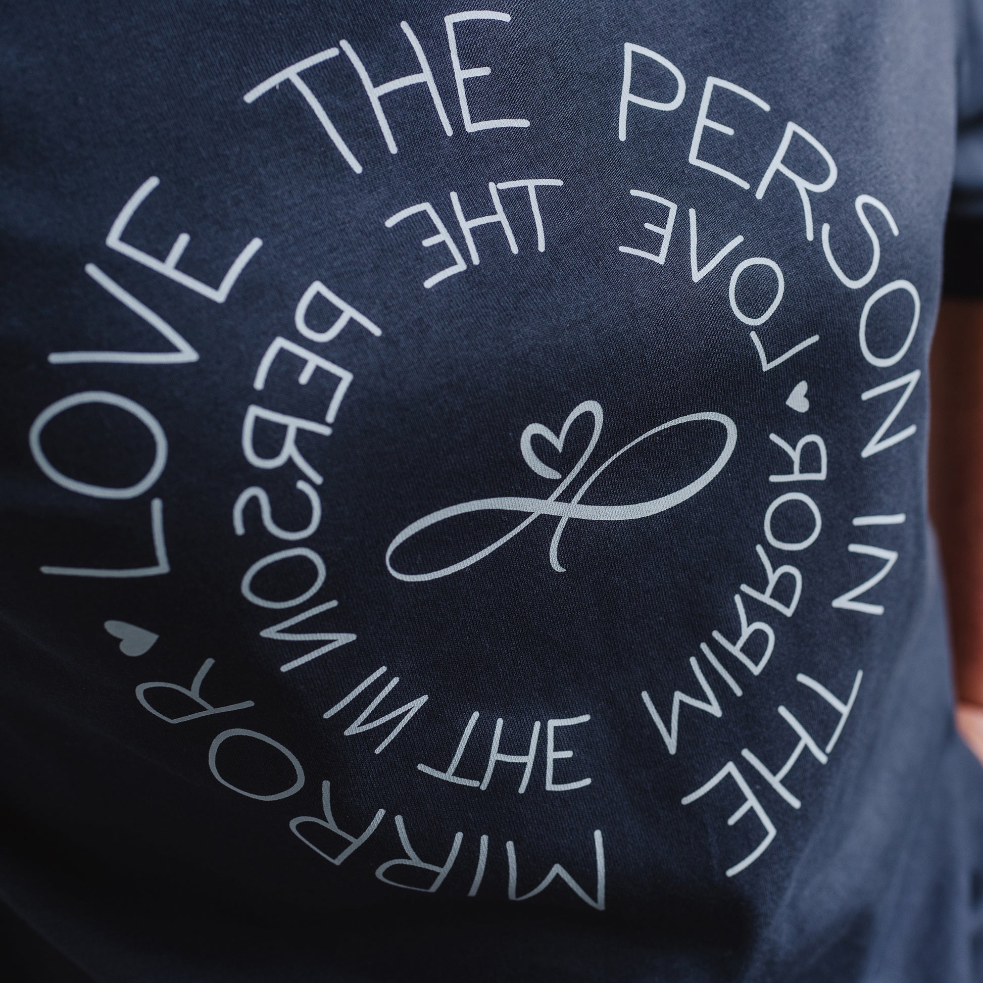 Women's Love the Person in the Mirror relaxed fit ink grey graphic t shirt with short sleeves. Infinity symbol and heart design and circular wording in white Love the Person in the Mirror in reverse on inner circle and right way round on outer circle so that wording.