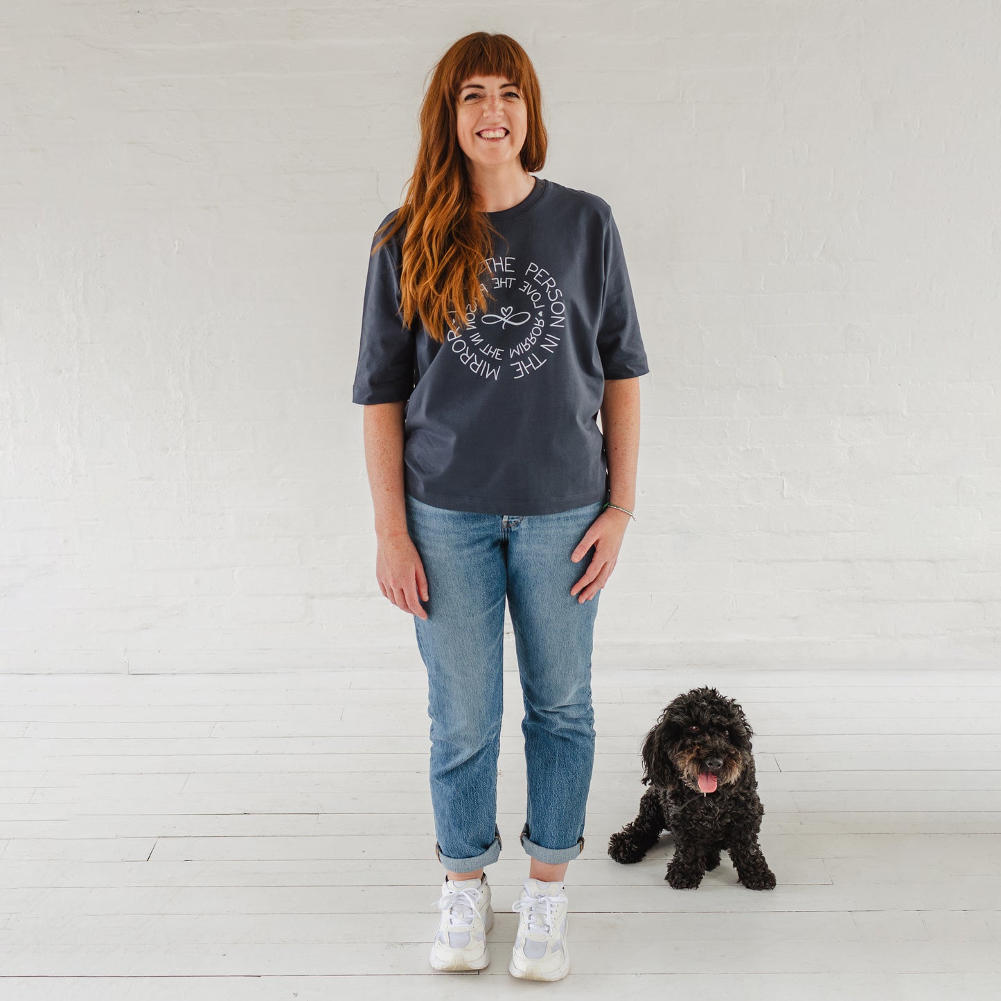 Auburn haired Caucasian woman wearing Love the Person in the Mirror women's relaxed fit ink grey slogan short sleeve boxy tee. Infinity symbol and heart design and circular wording in white Love the Person in the Mirror in reverse on inner circle and right way round on outer circle so that wording. Mid length sleeve to just above elbows. Woman wearing blue Jeans and white trainers. Cute cockapoo dog sitting next to her with his tongue out
