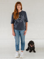 Auburn haired Caucasian woman wearing Love the Person in the Mirror women's relaxed fit ink grey slogan short sleeve boxy tee. Infinity symbol and heart design and circular wording in white Love the Person in the Mirror in reverse on inner circle and right way round on outer circle so that wording. Mid length sleeve to just above elbows. Woman wearing blue Jeans and white trainers. Cute cockapoo dog sitting next to her with his tongue out