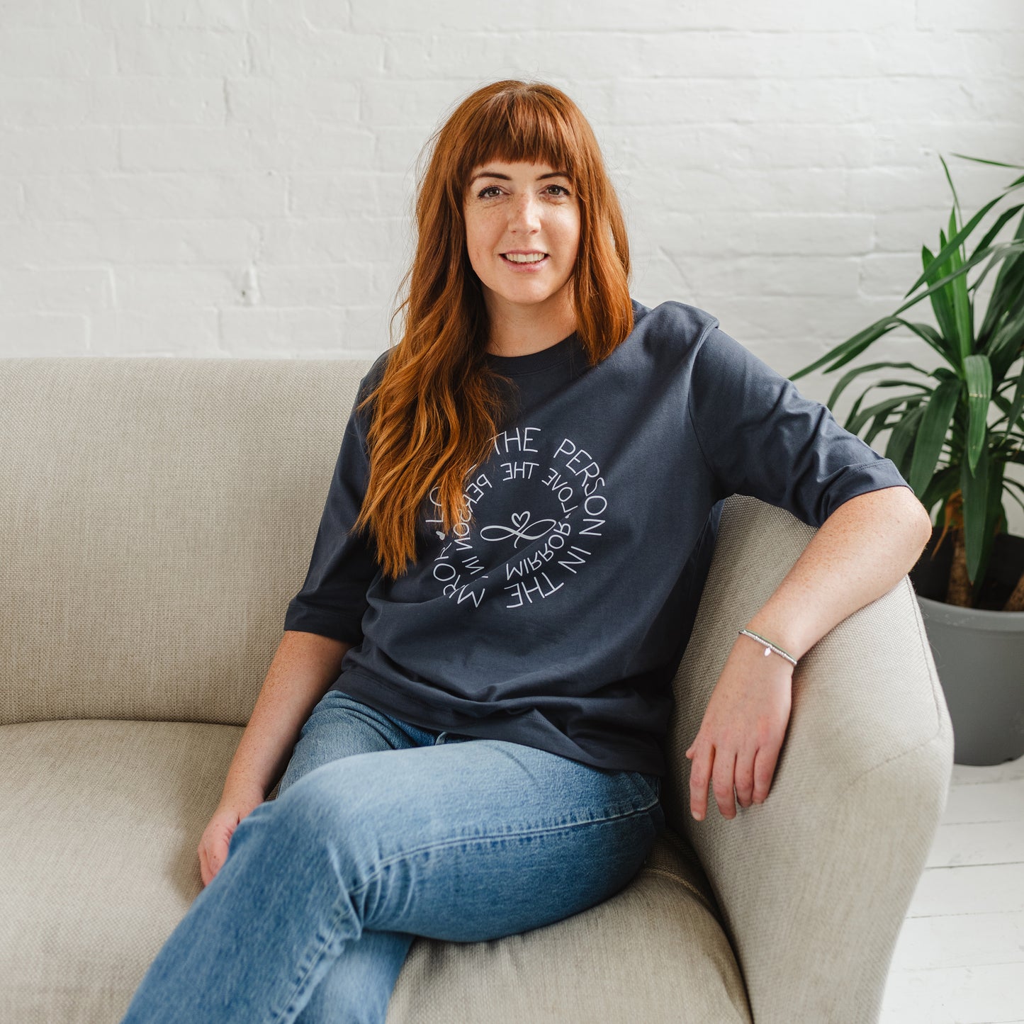 Caucasian woman sitting on sofa  wearing Women's Love the Person in the Mirror relaxed fit ink grey graphic t shirt with short sleeves. Infinity symbol and heart design and circular wording in white  Love the Person in the Mirror in reverse on inner circle and right way round on outer circle so that wording. Mid length sleeve to just above elbows. Woman wearing blue Jeans