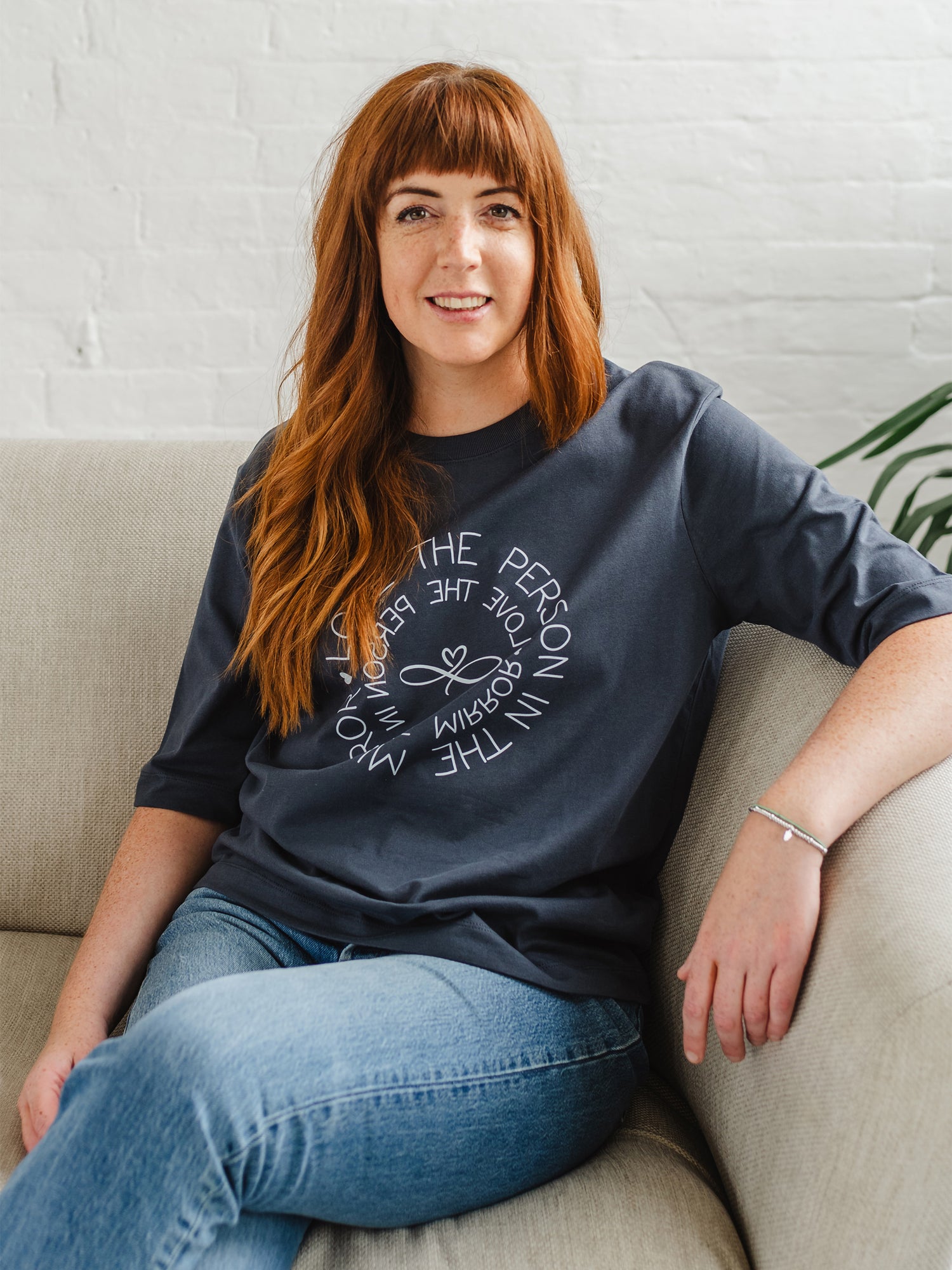 Caucasian woman sitting on sofa  wearing Women's Love the Person in the Mirror relaxed fit ink grey graphic t shirt with short sleeves. Infinity symbol and heart design and circular wording in white  Love the Person in the Mirror in reverse on inner circle and right way round on outer circle so that wording. Mid length sleeve to just above elbows. Woman wearing blue Jeans