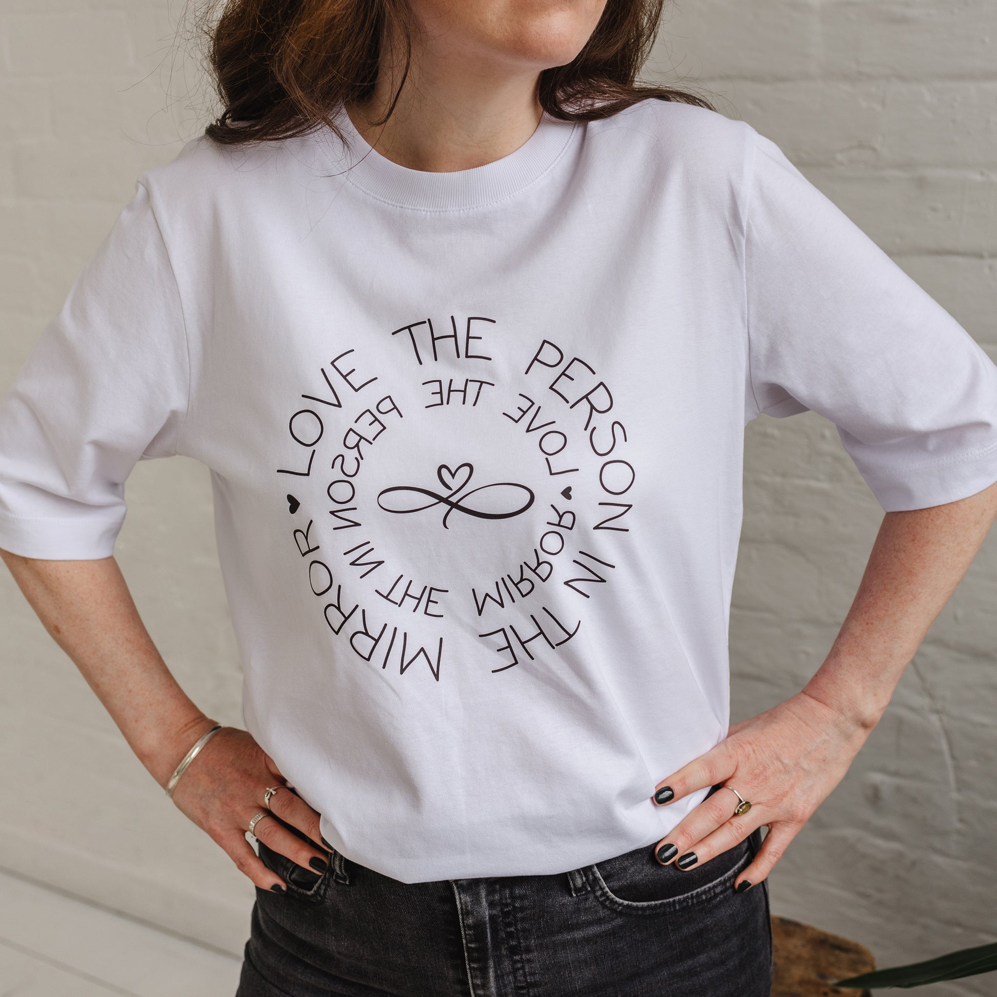 Woman wearing ladies white graphic t-shirt top. Design on fashion Tee reads Love the Person in the Mirror women's relaxed fit white slogan short sleeve boxy oversized t-shirt. Infinity symbol and heart design and circular wording Love the Person in the Mirror in reverse on inner circle and right way round on outer circle so that wording. Mid length sleeve to just above elbows. Worn with black jeans