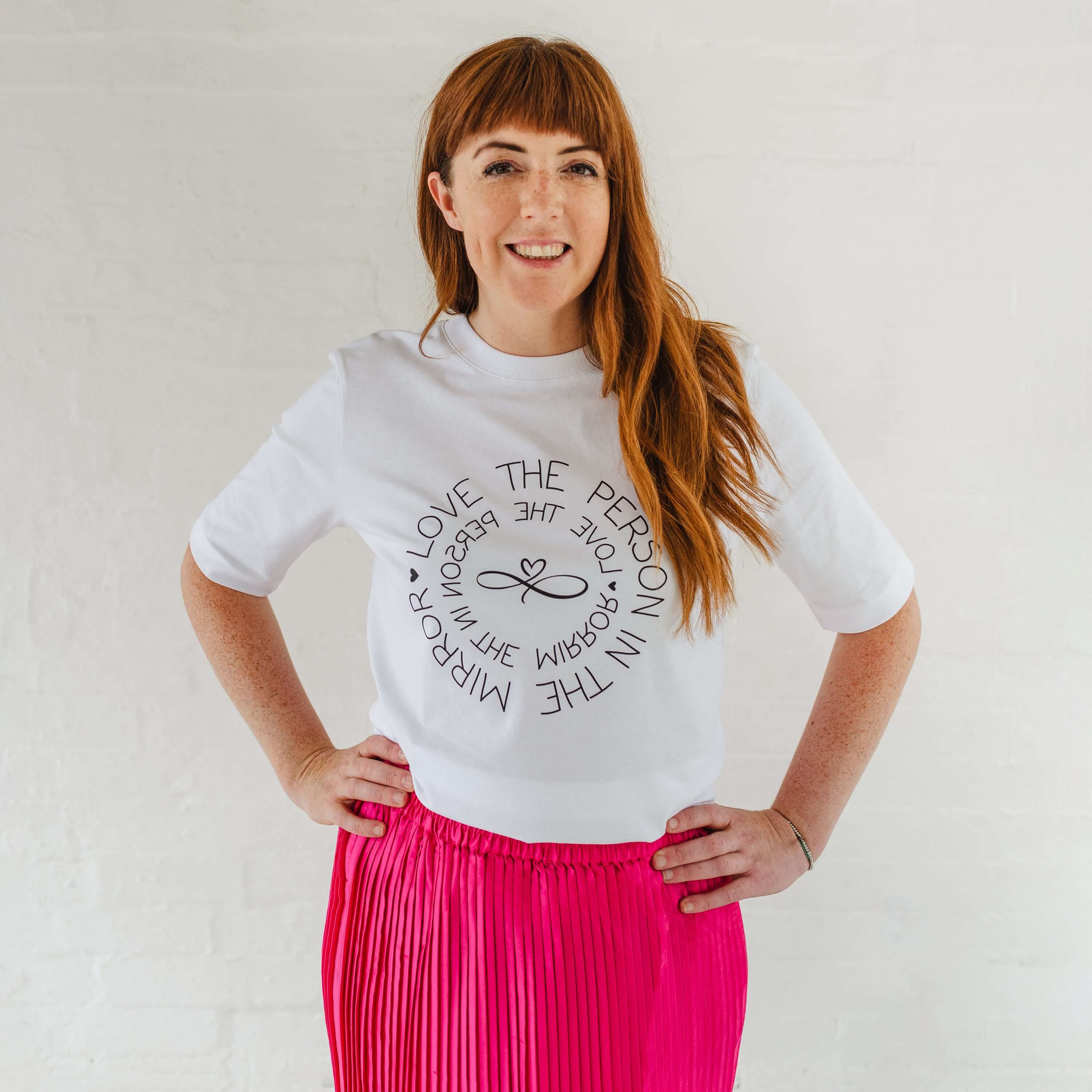 Caucasian woman wearing ladies white graphic t-shirt top. Design on fashion Tee reads Love the Person in the Mirror women's relaxed fit white slogan short sleeve boxy graphic tee. Infinity symbol and heart design and circular wording Love the Person in the Mirror in reverse on inner circle and right way round on outer circle so that wording. Mid length sleeve to just above elbows. Woman wearing bright pink mid length pleated fashion skirt