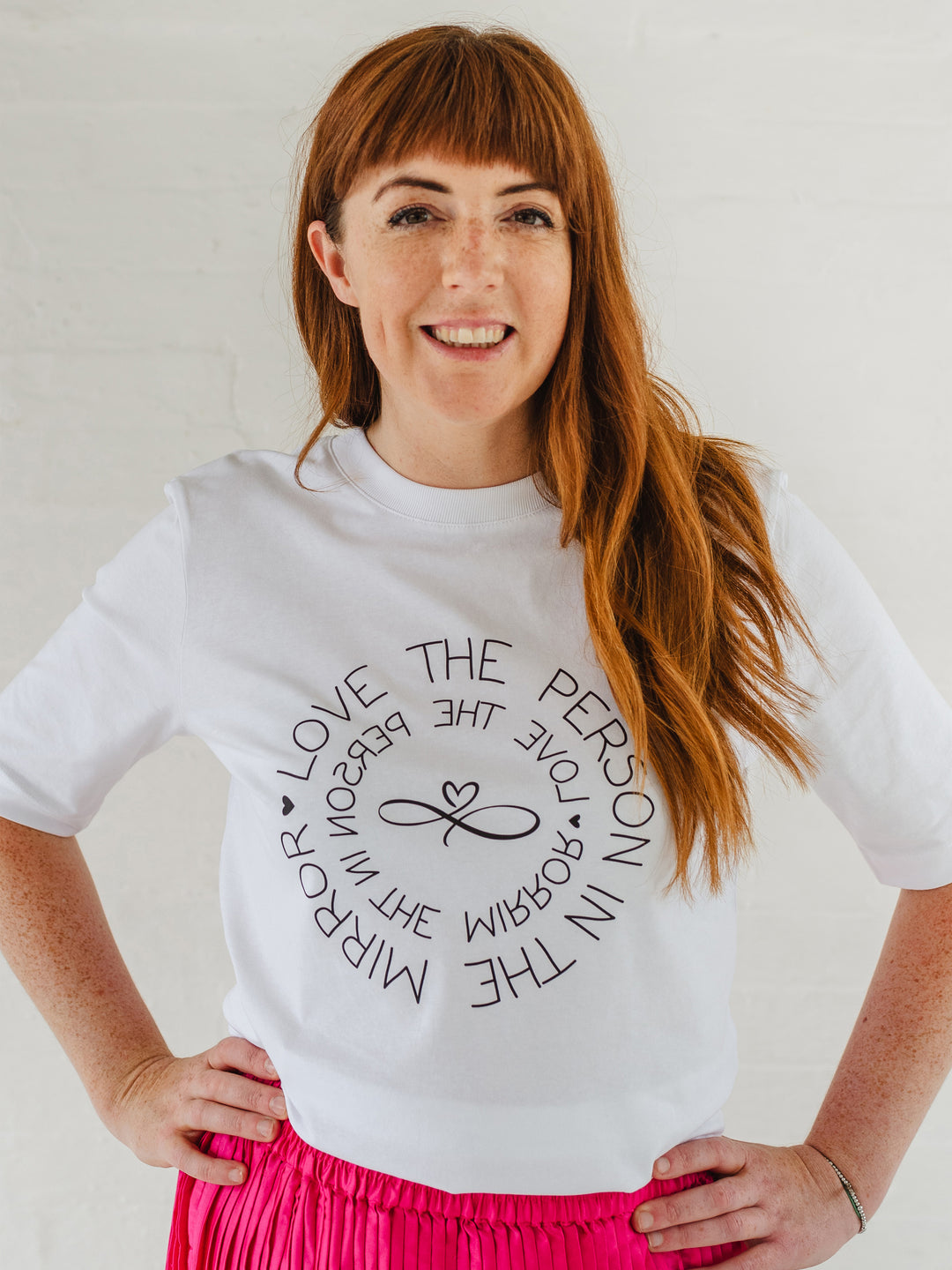 Caucasian woman wearing ladies white graphic t-shirt top. Design on fashion Tee reads Love the Person in the Mirror women's relaxed fit white slogan short sleeve boxy graphic tee. Infinity symbol and heart design and circular wording Love the Person in the Mirror in reverse on inner circle and right way round on outer circle so that wording. Mid length sleeve to just above elbows. Woman wearing bright pink mid length pleated fashion skirt