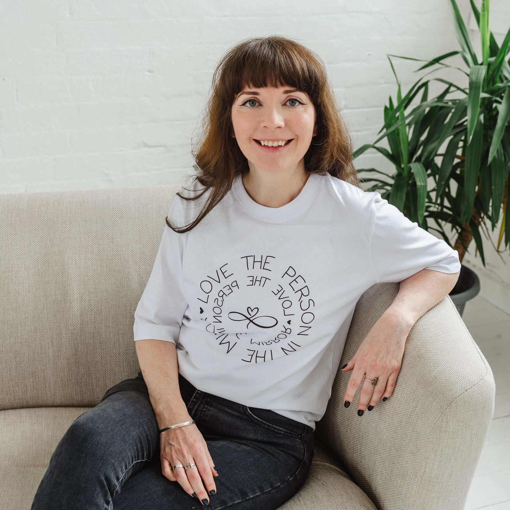 Woman wearing women's white graphic t-shirt top. Design on fashion Tee reads Love the Person in the Mirror women's relaxed fit white slogan short sleeve boxy graphic tee. Infinity symbol and heart design and circular wording Love the Person in the Mirror in reverse on inner circle and right way round on outer circle so that wording. Mid length sleeve to just above elbows. Woman is seated on a sofa, also wears black jeans.