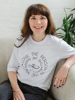 Woman wearing women's white graphic t-shirt top. Design on fashion Tee reads Love the Person in the Mirror women's relaxed fit white slogan short sleeve boxy graphic tee. Infinity symbol and heart design and circular wording Love the Person in the Mirror in reverse on inner circle and right way round on outer circle so that wording. Mid length sleeve to just above elbows. Woman is seated on a sofa, also wears black jeans.