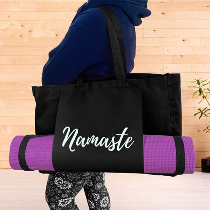 Woman wearing a navy hoodie with a black yoga mat bag over her shoulder. The bag has a yoga mat held long ways in the front pouch pockets and the words Namaste in silver across the front of the pouch. Woman is wearing black and white leggings. Yoga studio background
