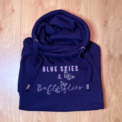 Casual Fashion Navy blue hoodie with cross cowl neck. Thumb holes to sleeves. Design is of four light grey butteflies in flight and wording Blue Skies and Butterflies to the front. Folded on a wooden floor. Unisex relaxed fit hoodie. Light purple wording with light grey back shadow. Long sleeves and straps from cowl neck with metal toggles on the end.  Blue skies text is all upper case. Butterflies text is handwritten style font upper and lower case