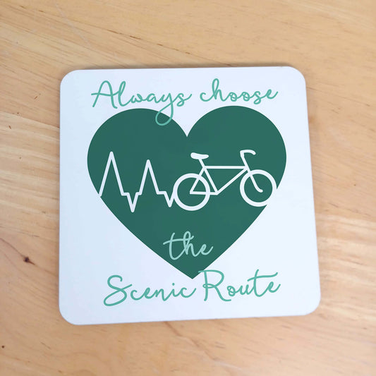 Gift Coaster for Cycling Enthusiast. White square wooden coaster with rounded edges. Green Heart with white heartbeat across turning into white bicycle outline. Cyclist theme wording Always choose the Scenic Route in dusky green handwriting style font