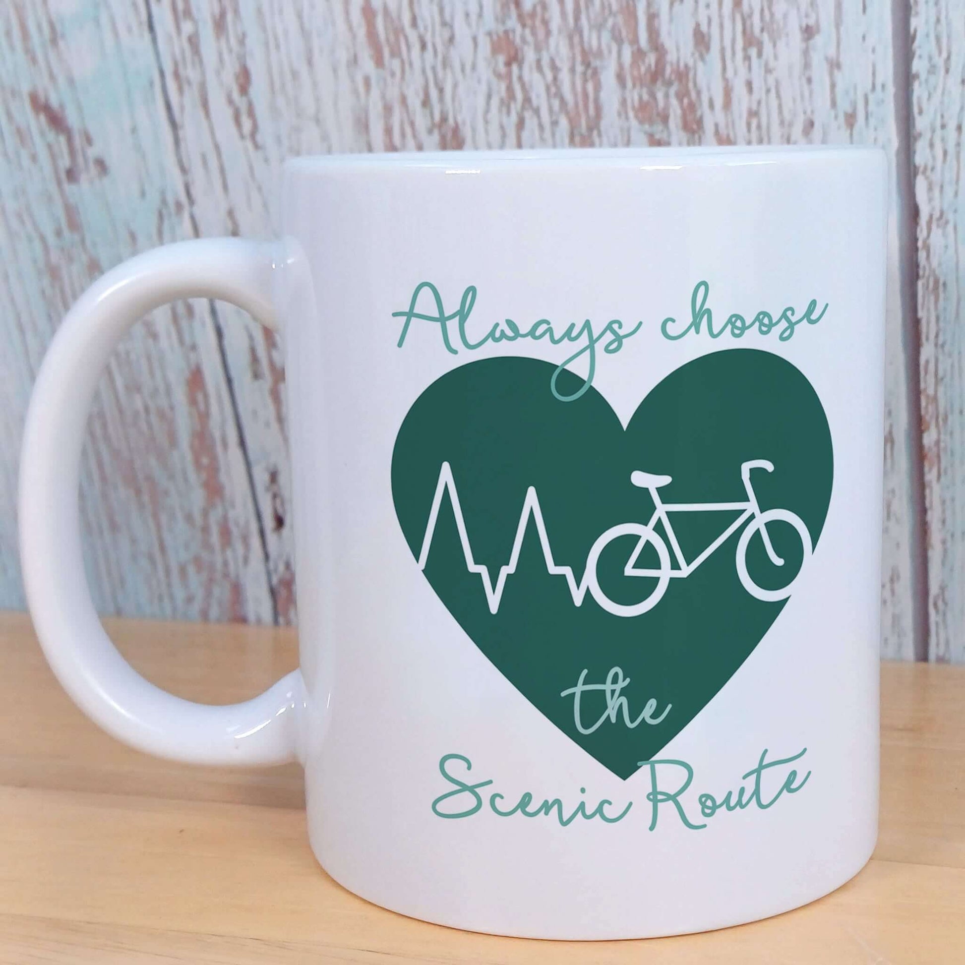 White Ceramic Gift Mug for Cycling Enthusiast. Cyclist theme Green Heart with white heartbeat across turning into white bicycle outline. Keen cyclist theme wording Always choose the Scenic Route in dark green handwriting style font