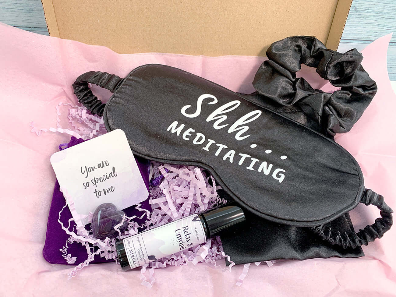 Brown gift box with pink tissue paper inside is a satin black sleep mask with 'Shh...meditating' matching hair scrunchie, essential oil roll on and purple amethyst keepsake with card 'You are so special to me' 