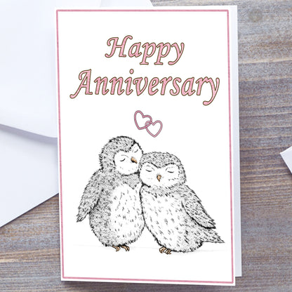 White gloss A5 Happy Anniversary Greeting card with pink text and border. Optional name personalisation. Two hand-drawn fluffy grey owls snuggling under interlocking pale pink hearts. Happy Anniversary in Pink at top of card. Non-personalised version. Wedding Anniversary Card, white envelope