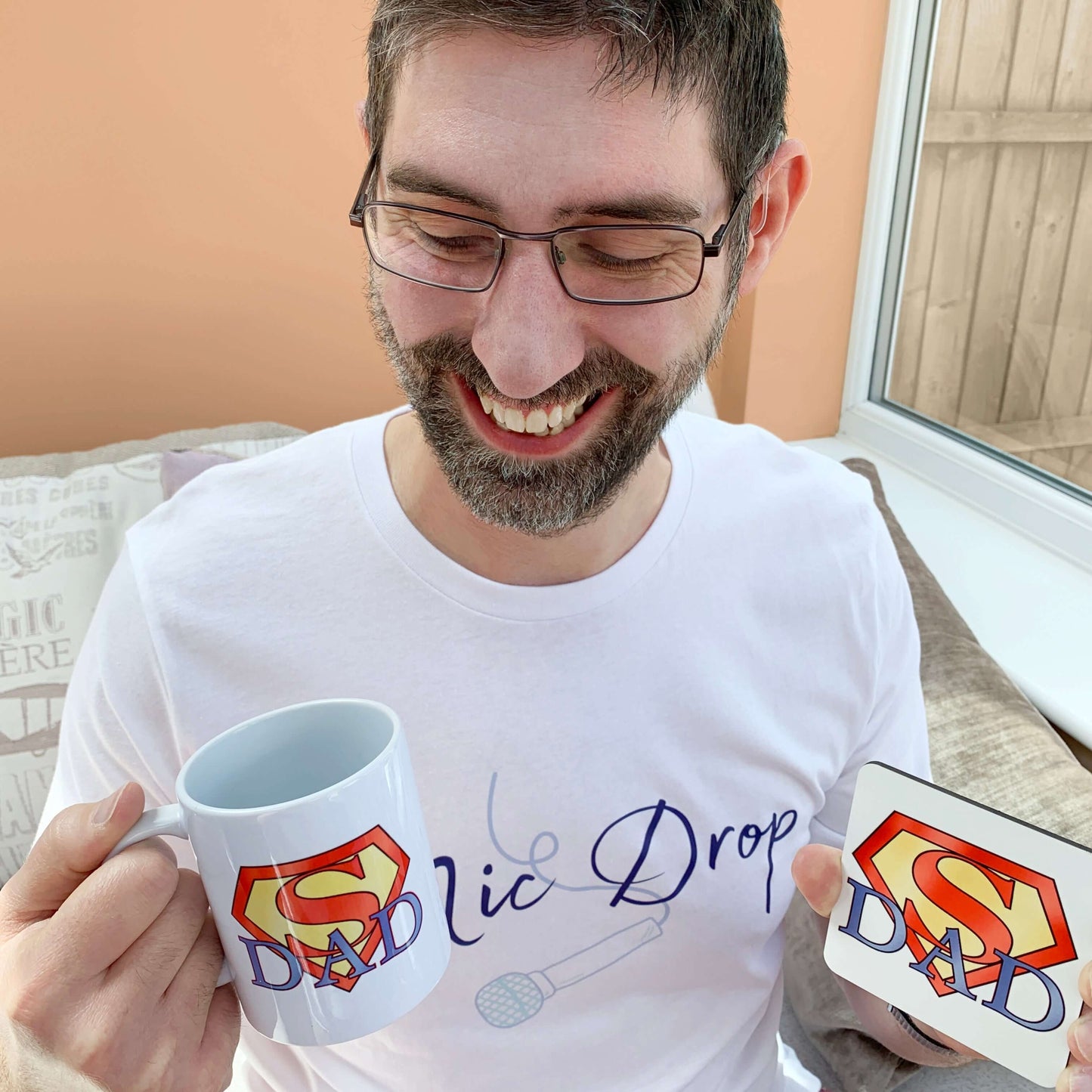 Personalised gift for dad. Daddy smiling looking down at his present of a personalised Super Dad Mug and matching Superhero themed coaster. Superman Logo design with Dad intertwined underneath. Personalised message on reverse side of mug unseen in image