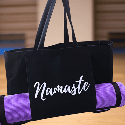 Gift for Yoga Lovers, a Yoga Mat bag with Silver script style writing Namaste across a front pouch pocket. Large black tote bag with shoulder straps. Lavender purple yoga mat held long ways rolled up in straps in the pouch pocket. Background of a yoga studio out of focus