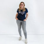 Woman standing smiling has long blonde hair and wears trendy Womens Leopard Print Fashion Top. Navy blue t-shirt with round neck and short sleeves. Organic cotton ladies t shirt with multi colour spots animal print slogan on Tee reads WILD, worn with grey skinny jeans and trainers. 