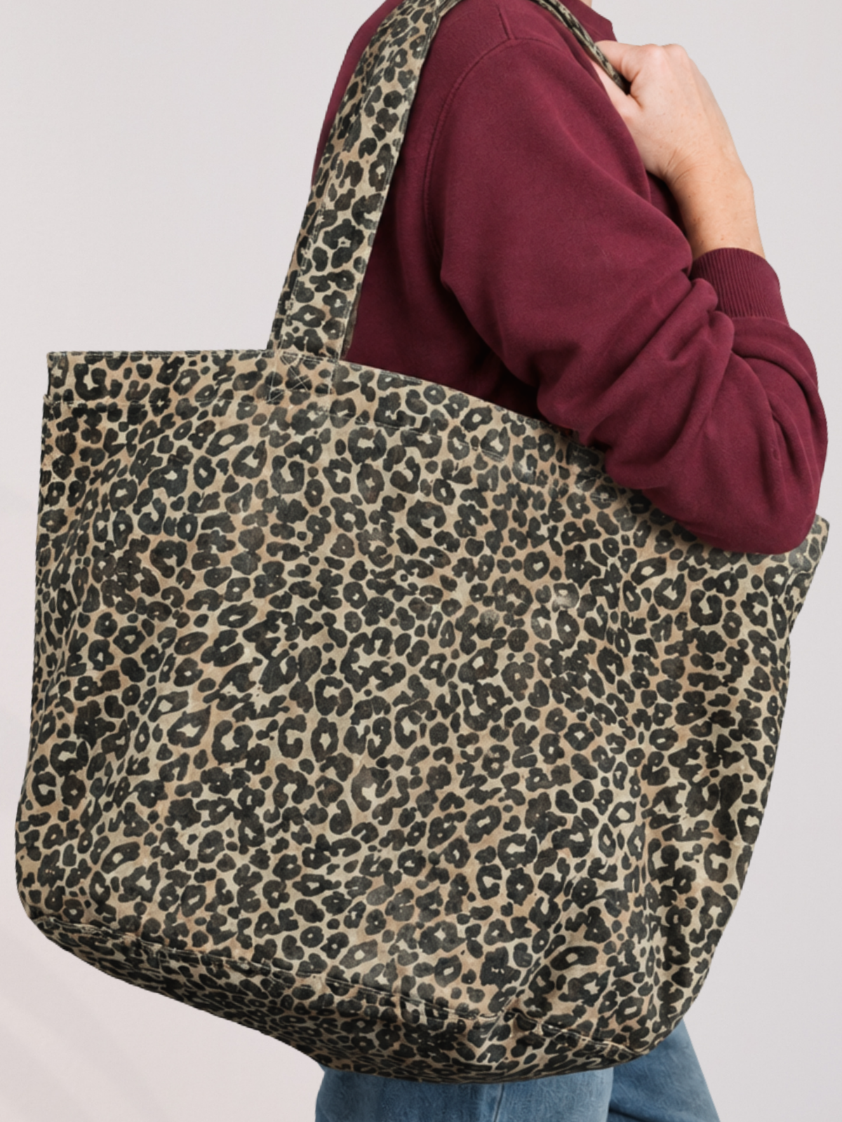 A woman wearing a burgundy sweatshirt and blue jeans carries a large canvas tote bag with a washed effect beige, brown, and black leopard print pattern against a plain light background.