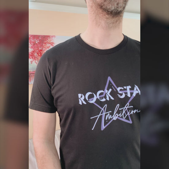 Video of Man wearing mens black short sleeve graphic tshirt. Crew neck casual wear. Band Tee Design is mid purple star outline behind wording ROCK STAR in upper case shadowed  light grey/purple text, then Ambition below in script. Rock music themed unisex top