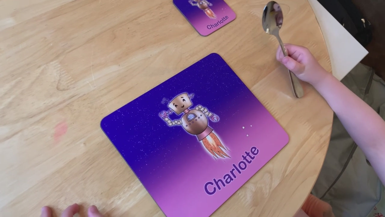 Video panning close up of Pink and purple star sky kids placemat set. Robot smiling with jets flying into space. Personalised with girl's name at the bottom of custom child's dinner mat and matching coaster.  Childs hand in shot at a table holding Kids spoon 
