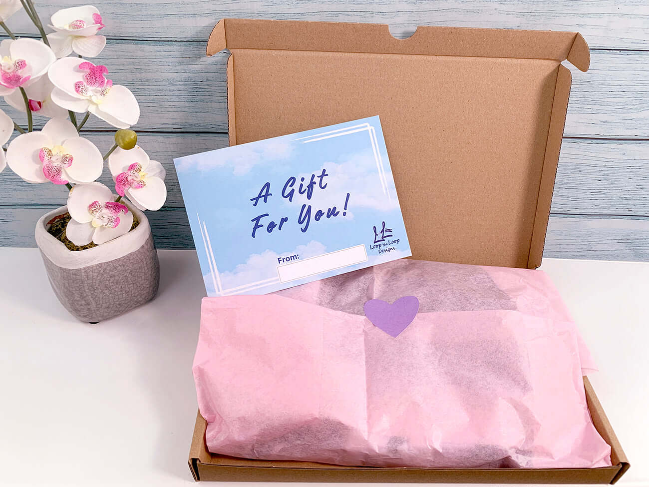 Gift box - cardboard with pink recycled tissue paper gift wrap and heart sticker closure. Gift insert card that has blue sky and clouds reads A Gift for You! 