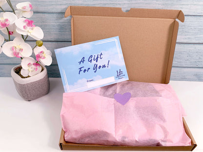 Gift wrapped with pink tissue paper and purple heart sticker to Close. Gift slip sky and cloud background design with wording A Gift for You and blank white box to add from: ....... Loop the Loop Designs logo bottom right. Pink orchid faux plant to right of giftset box