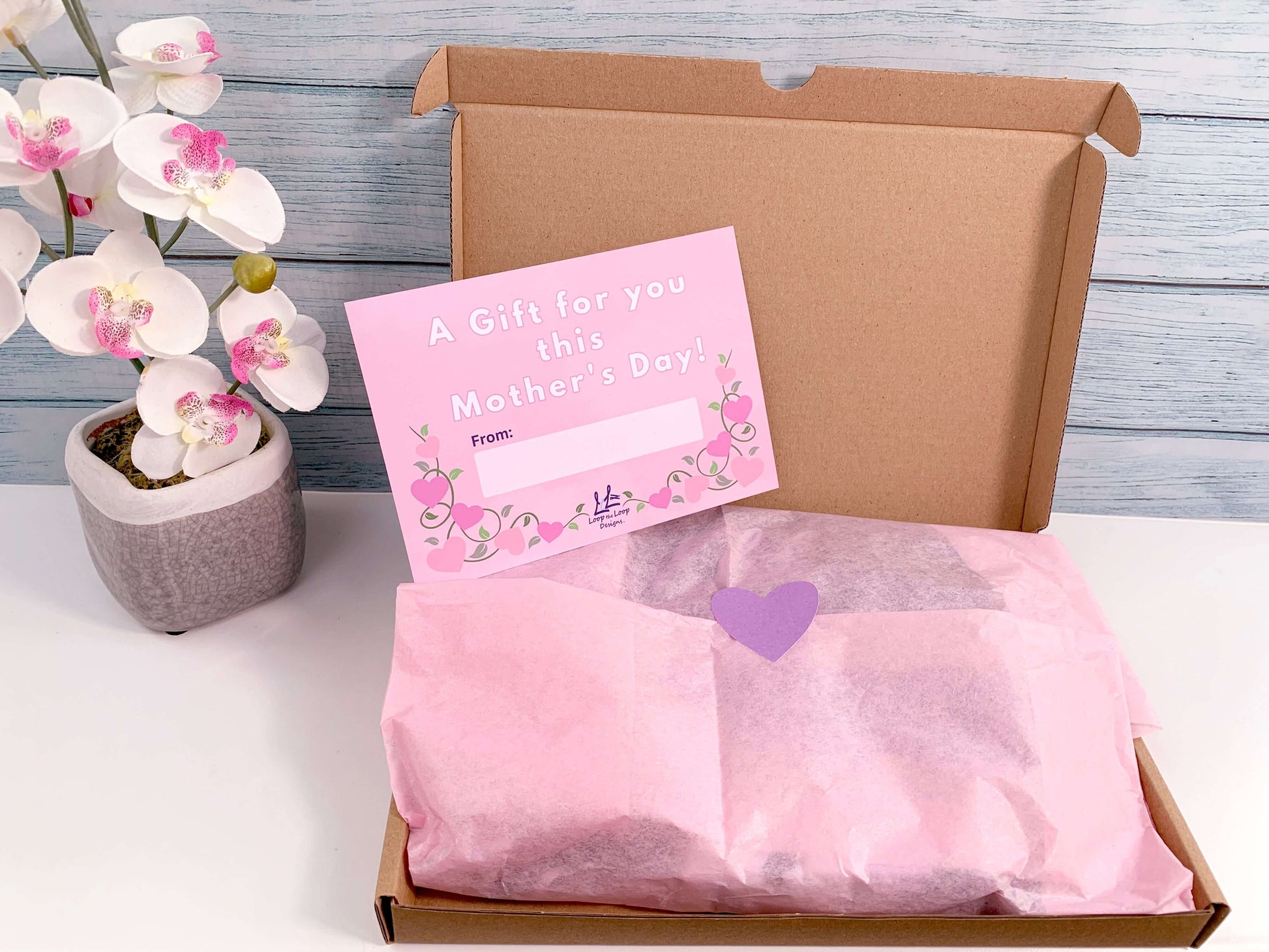 Gift wrapped with pink tissue paper and purple heart sticker to Close. Gift slip pink background design with wording A gift for you this Mothers Day heart shape pink flowers ona vine below, and blank white box to add from: ....... Loop the Loop Designs logo bottom centre. Pink orchid faux plant to right of giftset box
