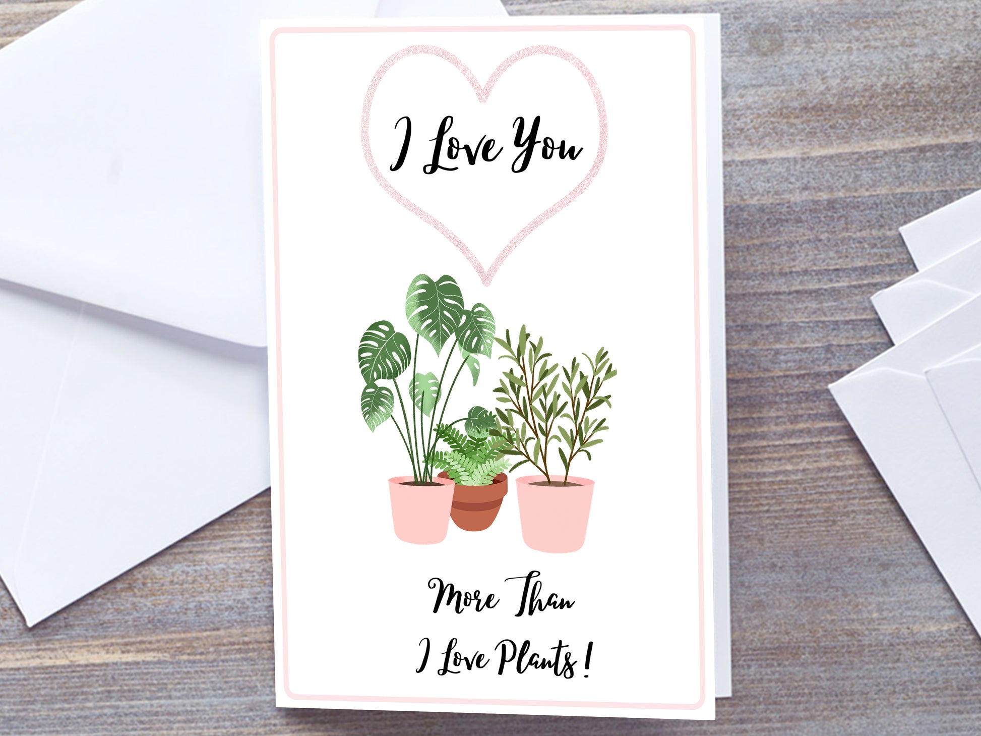 Plant lover Greetings card for anniversary or valentines. White card to 3 House plants in pink plant pots. Wording I love you inside a pink heart outline. Below plant images is wording More than I love plants! In handwriting script text. Card resting on Grey wooden background with white envelope 