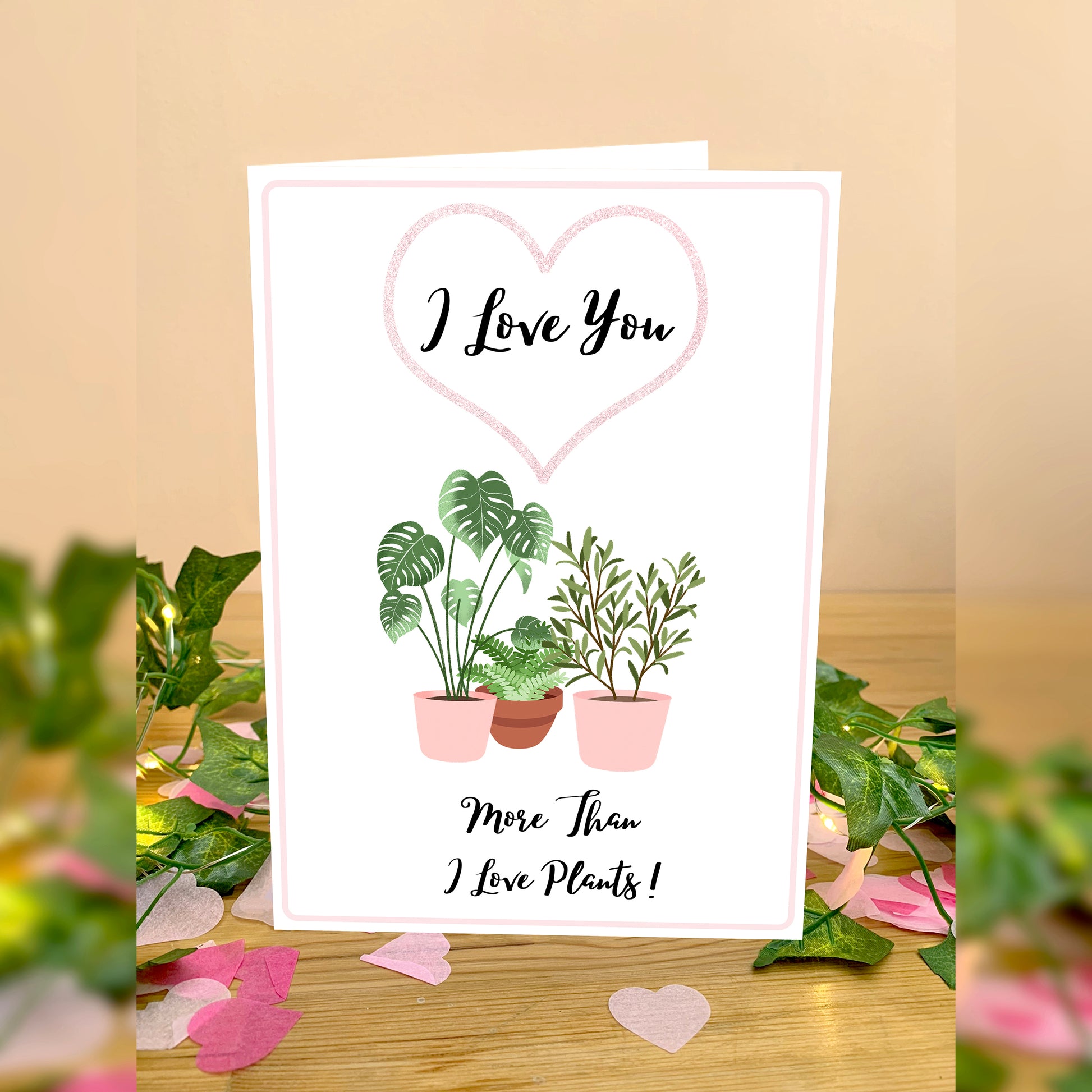 Plant lover Greetings card for anniversary or valentines. White card to 3 House plants in pink plant pots. Wording I love you inside a pink heart outline. Below plant images is wording More than I love plants! In handwriting script text. Flower petals scattered around card placed on a wooden table with leaves on