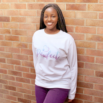 Afro-caribbean woman wearing white relaxed fit long sleeve sweatshir. Two tone blue fading to dark pink Slogan sweater design wording Pawfect same two-tone colour heart outline above with dog paw print within outline. Woman wears rust colour jeans and is stood in outdoors in front of a brick wall, Model wears purple jeans with the top and is smiling