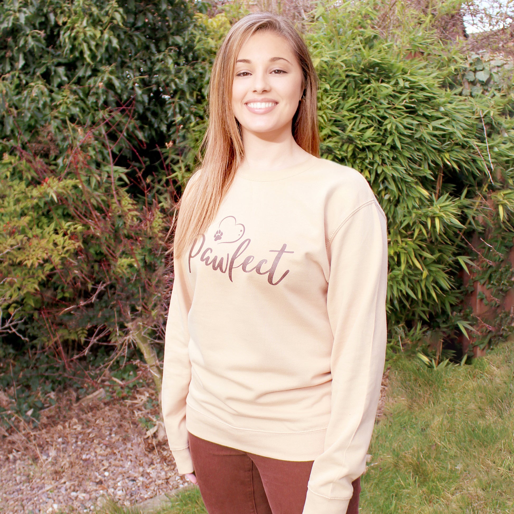 Beige / Caramel colour relaxed fit long sleeve sweatshirt worn by caucasian woman with long brown hair. Slogan sweater design wording Pawfect in brown font and brown heart outline above with dog paw print within outline. Woman wears rust colour jeans and is stood in outdoors in a garden with plants behind