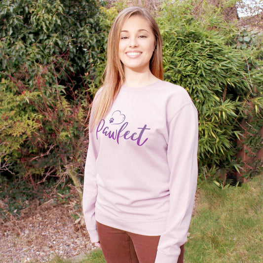 Light pink relaxed fit long sleeve sweatshirt worn by caucasian woman with long brown hair. Slogan sweater design wording Pawfect in purple font and purple heart outline above with dog paw print within outline. Woman wears rust colour jeans and is stood in outdoors in a garden with plants behind