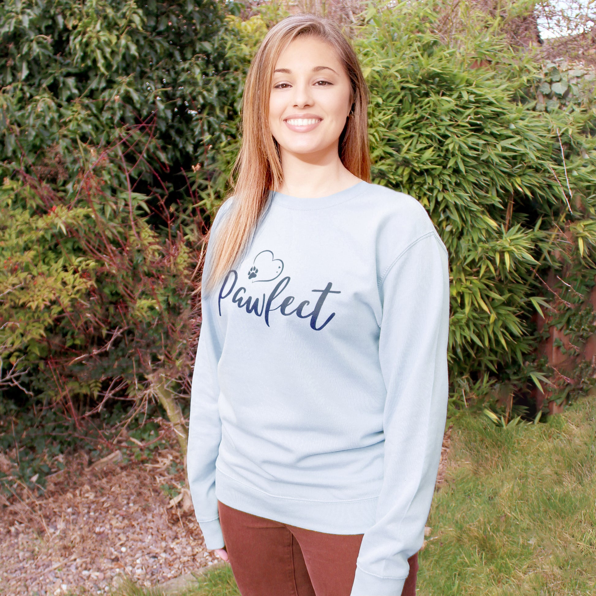 Light blue relaxed fit long sleeve sweatshirt worn by caucasian woman with long brown hair. Slogan sweater design wording Pawfect in dark blue font and navy blue grey heart outline above with dog paw print within outline. Woman wears rust colour jeans and is stood in outdoors in a garden with plants behind