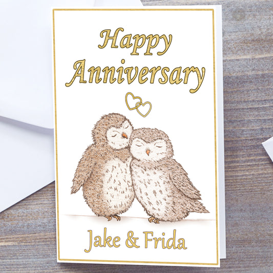White card with gold border. Gloss A5 Happy Anniversary Greetings Card. Owl and heart design. two hand-drawn fluffy owls snuggling under interlocking gold hearts. Personalised with recipients name(s) in gold text below.  Happy Anniversary wording above interlocking hearts in Gold font. Wedding Anniversary Card, white envelope