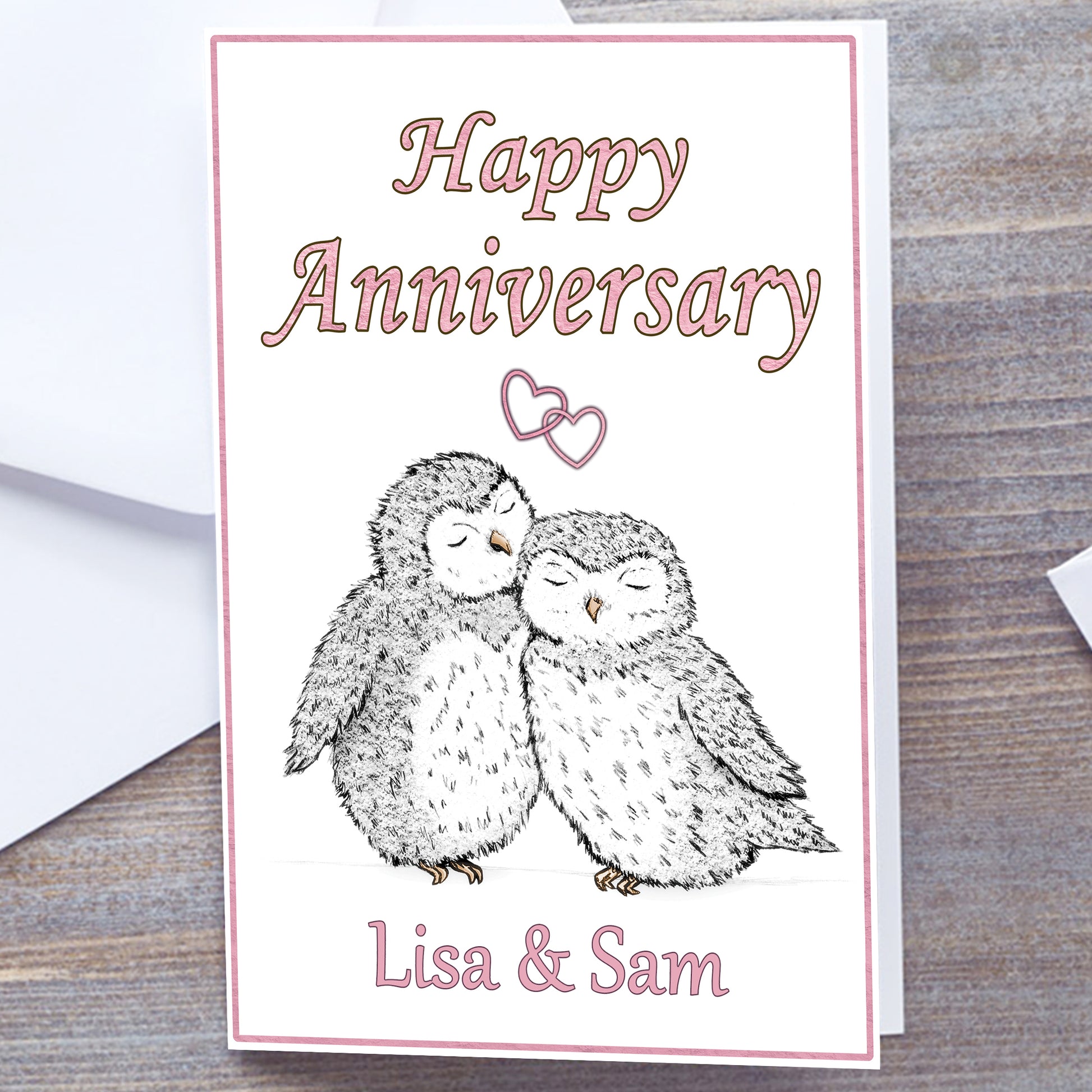 White card with pink border. Gloss A5 Happy Anniversary Greeting Card. Owl and heart design. Two black and white hand-drawn fluffy owls with pale yellow beaks and feet snuggling under interlocking pink hearts. Personalised with recipients name(s) in pink text below. Happy Anniversary wording above interlocking hearts in pink font. Wedding Anniversary Card, white envelope
