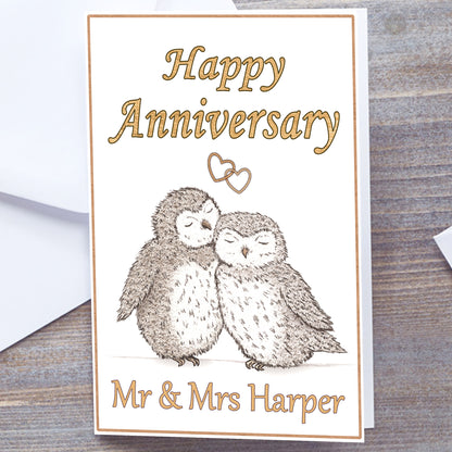 Personalised White gloss A5 Happy Anniversary Greeting card with gold text and border.  Grey-Brown Owls. Two hand-drawn fluffy owls snuggling under interlocking gold hearts. Happy Anniversary in Gold at the top. Recipients name(s) in gold at bottom. Wedding Anniversary Card, white envelope