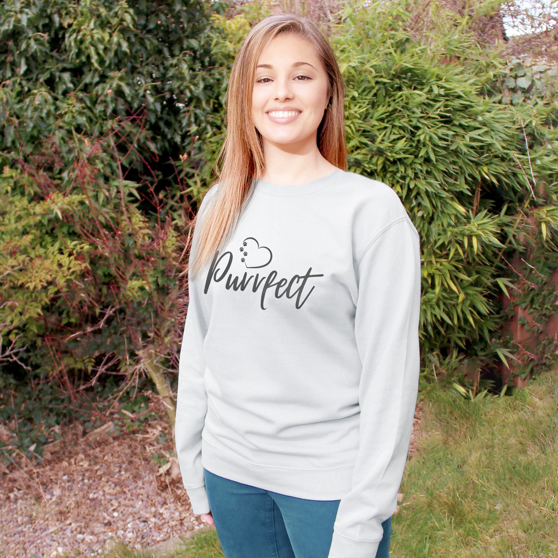 Light grey relaxed fit long sleeve sweatshirt worn by caucasian woman with long brown hair. Slogan sweater design wording Purrfect in dark grey font and charcoal grey heart outline above with small cat paw prints within outline. Woman wears teal colour jeans and is stood in outdoors in a garden with plants behind