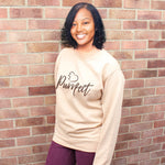 Cat lover top - Beige / Caramel colour relaxed fit long sleeve sweatshirt. Slogan sweater design wording Purrfect in brown font and brown heart outline above with cat 4 sequenced paw print within outline. Model stands outdoors against a brick wall smiling wearing plum jeans. She has dark skin and kid length curled hair