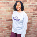 Cat lover top - Light sky blue colour relaxed fit long sleeve sweatshirt. Slogan sweater design wording Purrfect in dark blue font and  navy heart outline above with cat 4 sequenced paw print within outline. Model stands outdoors against a brick wall smiling wearing plum jeans. She has dark skin and mid length curled hair