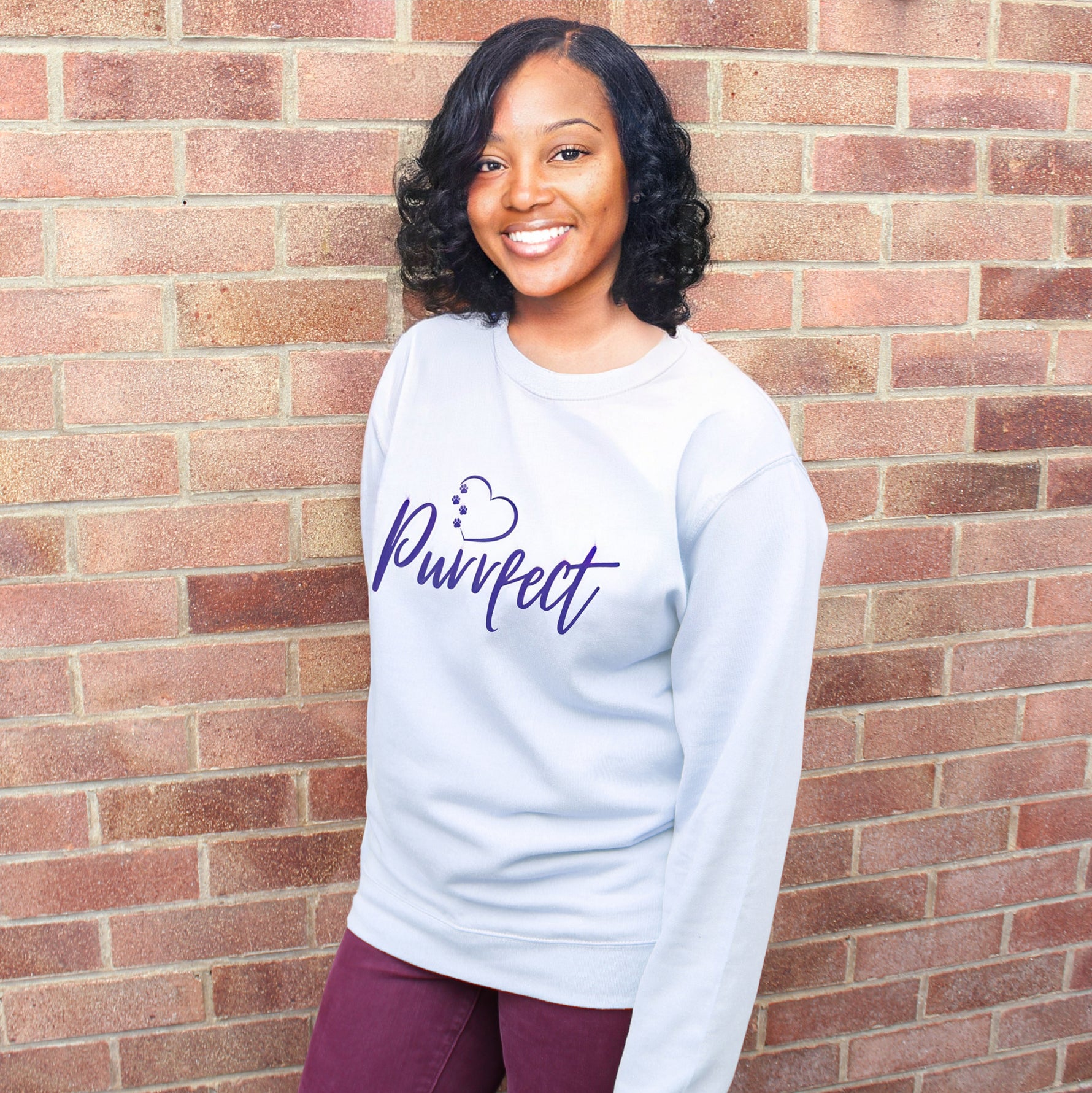 Cat lover top - Light sky blue colour relaxed fit long sleeve sweatshirt. Slogan sweater design wording Purrfect in dark blue font and  navy heart outline above with cat 4 sequenced paw print within outline. Model stands outdoors against a brick wall smiling wearing plum jeans. She has dark skin and mid length curled hair