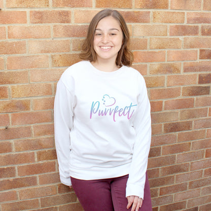 White top - relaxed fit long sleeve sweatshirt worn by caucasian woman with mid length brown hair. Slogan sweater design wording Purrfect in two-tone blue-purple heart outline above with 4 sequenced cat paw prints within outline. Woman wears plum colour jeans and is stood in outdoors in front of brick wall