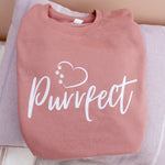 Cat mum top folded resting on a cushion - Dusky pink colour relaxed fit long sleeve sweatshirt on wooden hanger Slogan sweater design wording Pawfect in white font and brown heart outline above with cat 4 sequenced paw print within outline.