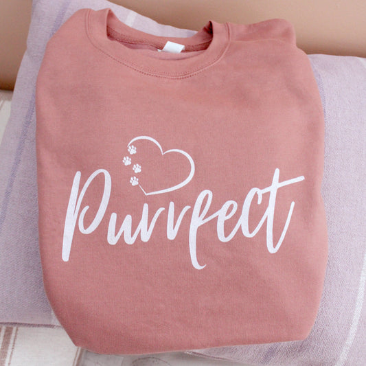Cat mum top folded resting on a cushion - Dusky pink colour relaxed fit long sleeve sweatshirt on wooden hanger Slogan sweater design wording Pawfect in white font and brown heart outline above with cat 4 sequenced paw print within outline.